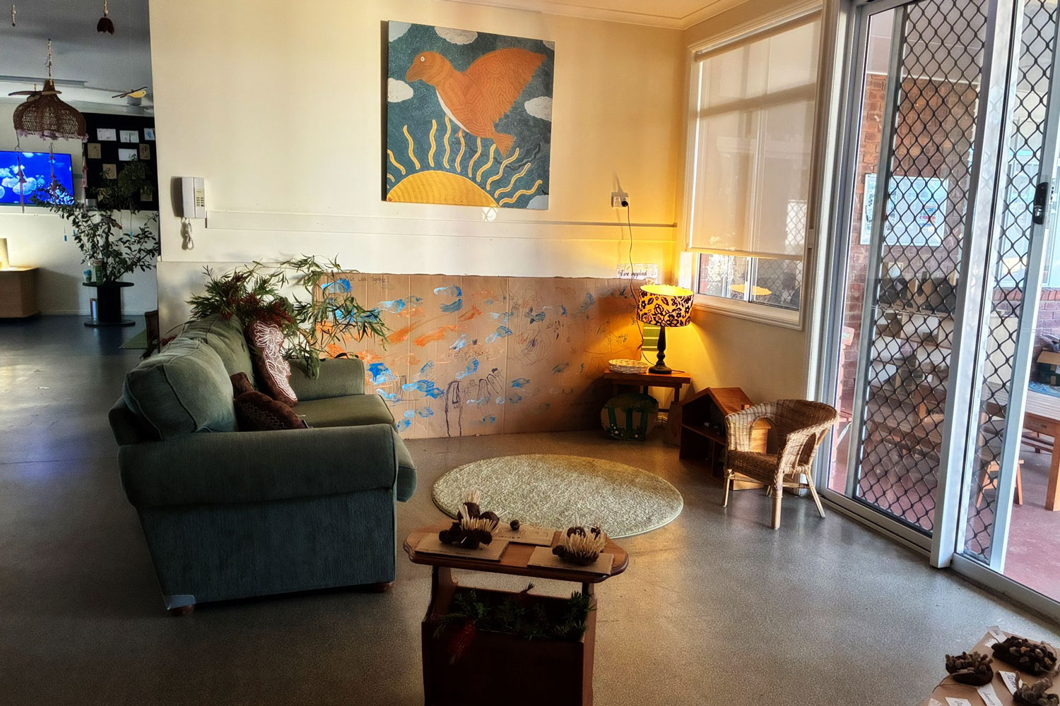 A photograph of an indoor kindergarten space. There is a green couch, a small wicker chair, a rug and a lamp on. The walls are adorned with children&rsquo;s artwork