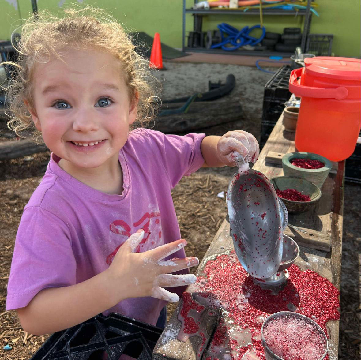 A photograph of a child playing outside with red liquid in a different containers.